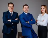 Trzy osoby w garniturach i białej koszuli, stojące na szarym tle, z rękami skrzyżowanymi na piersi.