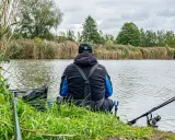 Wędkarz siedzi nad rzeką, przygotowując zanętę na rzekę feeder. W tle trzciny i drzewa.