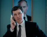 Un uomo in giacca e cravatta parla al telefono, con un ritratto di Putin sullo sfondo. Sembra il mago del Cremlino.