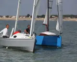 Deux voiliers naviguent côte à côte. Sur le premier, un homme en blanc manœuvre. Le second, d'un bleu vif, est un aloes 18.