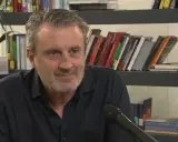 Uomo con barba grigia sorride, microfono davanti. Alle sue spalle scaffali pieni di libri, tra cui "Le ossa parlano trama".