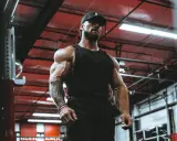 Ein Mann mit beeindruckendem **mens physique** steht im Fitnessstudio. Seine Muskeln sind stark definiert, er trägt eine schwarze Kappe und ein Tanktop.