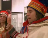 Två personer i samiska dräkter, en kvinna med röd mössa och en man med färgglad mössa. De är same folk.