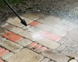 Czyszczenie kostki brukowej myjką ciśnieniową. Woda usuwa brud z czerwonych i szarych płyt.