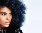 Mujer con cabello rizado negro azulado y maquillaje vibrante, luciendo una chaqueta brillante.