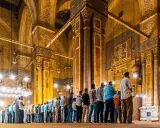 Männer beten Tarawih in einer Moschee. Sie stehen in Reihen vor kunstvoll verzierten Säulen und Bögen, beleuchtet von zahlreichen Lampen.