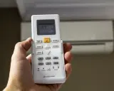 Mano sujetando mando para configurar mando universal aire acondicionado sin código. Aire acondicionado al fondo.