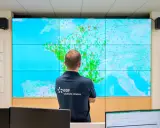 Un homme regarde une carte de la France sur un grand écran, avec des points verts et rouges. Il porte un polo "EDF solutions solaires", peut-être pour surveiller les installations et éviter une arnaque panneau solaire.