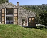 Una casa in pietra con grandi porte in legno, perfetta per chi ama vivere in campagna. Si intravedono interni moderni e un paesaggio collinare rigoglioso.
