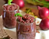 Dwa słoiczki z domowym, aromatycznym chutney z czerwonej cebuli, ozdobione listkiem zieleniny.