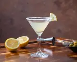 Orzeźwiający drink z martini, ozdobiony plasterkiem cytryny i solnym rantem. Obok leżą połówki cytryn, oliwki i akcesoria barmańskie. Idealny na domowe drinki z martini.