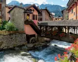 Un pittoresco ponte di legno attraversa un torrente impetuoso, fiancheggiato da case colorate e fiori rossi. Cosa vedere in questo incantevole borgo alpino.