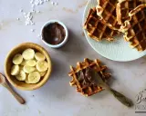 Une gaufre bubble est garnie de pâte à tartiner au chocolat. À côté, des rondelles de banane et une cuillère en bois.
