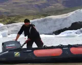 Un homme tente de démarrer un moteur 2 temps sur un Zodiac, mais il cale. Derrière lui, des icebergs flottent dans un lac glaciaire.