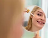 Mujer rubia sonríe mientras se aplica aceite facial con gotero en la mejilla. Un paso esencial en su rutina de cuidado de la piel, ya sea aceite facial antes o después de la crema.