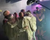 The ark medlemmar i vita, glittriga scenkläder omfamnar varandra medan en filmare spelar in.