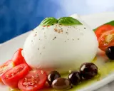 Duża kulka mozzarelli di bufala z pomidorkami koktajlowymi, oliwkami i bazylią na białym talerzu.