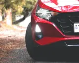 Czerwony Hyundai i20 z zapalonymi światłami dziennymi i reflektorami. Widać fragment maski, grill i tablicę rejestracyjną.