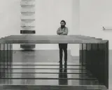 A man stands with arms crossed, contemplating a minimalist installation of Donald Judd art, featuring stacked shelves and a long, rectangular sculpture.