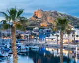Palme e barche nel porto di Alicante, con il Castello di Santa Barbara illuminato sullo sfondo.