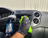 Ręce w czarnych rękawiczkach czyszczą deskę rozdzielczą samochodu, używając płynu "Wash Mist". To prosty sposób, jak pomalować plastiki w samochodzie, nadając im nowy wygląd.
