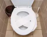 Inodoro blanco con papel higiénico cruzado sobre la tapa, listo para desatascar wc. Cerca, un cesto de basura y un cepillo.