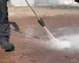 Osoba w roboczych spodniach i butach czyści mokrą nawierzchnię za pomocą myjki ciśnieniowej z dyszą rotacyjną.
