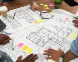 Des mains examinent un plan d'une maison, avec des post-it colorés et un téléphone sur la table.