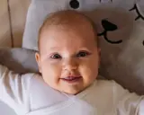 Uśmiechnięte niemowlę w białym body leży na miękkiej poduszce w kształcie misia. To mądre imię dla chłopca, które przyniesie mu szczęście.