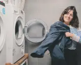 Kobieta z uśmiechem trzyma granatową kurtkę, przygotowując się do prania Gore-Tex. Obok stoi otwarta suszarka.