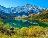 Widok na Morskie Oko z wysokości. Turkusowa tafla jeziora odbija ośnieżone szczyty Tatr. Jesienne drzewa otaczają brzeg.