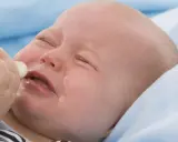 Ein weinendes Baby mit geschlossenen Augen, dem eine Flasche angeboten wird. Das Baby will nicht trinken.