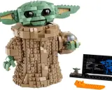 Figurka Grogu z klocków LEGO, znana z gier LEGO Star Wars, trzyma srebrną kulę. Obok stoi mała figurka i tabliczka informacyjna.