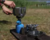 Na stole piknikowym stoi menażka, naczynie turystyczne, obok kubek i talerz.