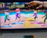 Ręka trzymająca kontroler do gry, na ekranie telewizora widać kolorowe postacie z gry ruchowej Nintendo Switch.