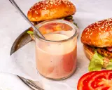 Ein Glas mit cremiger, rosafarbener beste burger sauce steht neben zwei saftigen Burgern. Ein Löffel ragt aus dem Glas.