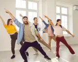 Grupa ludzi ćwiczy energiczne samba kroki w jasnej sali tanecznej, z rękami uniesionymi w rytm muzyki.