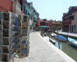 Cartoline di Murano, Burano e Torcello in vendita lungo un canale con case colorate e barche ormeggiate.