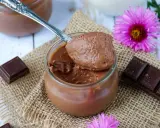 Une cuillère de crème au chocolat sans sucre, onctueuse et aérienne, sur un pot en verre. Des carrés de chocolat et des fleurs roses décorent la scène.