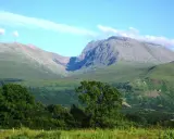 Majestatyczny Ben Nevis wznosi się na tle błękitnego nieba z delikatnymi chmurami. Zielone zbocza górskie i bujna roślinność tworzą malowniczy krajobraz.