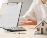 Une personne présente un contrat, une calculatrice, des clés et une maquette de maison. L'ambiance est un peu sinistre, comme si un problème était sur le point d'être révélé.