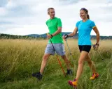 Ein Paar genießt das **power walking** in einer sonnigen Landschaft mit hohem Gras und bewaldeten Hügeln im Hintergrund.