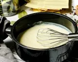 Cremige Béchamelsauce wird in einem Topf mit einem Schneebesen zubereitet. Daneben liegen Lasagneplatten und Gewürze.
