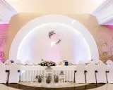 Elegancka sala weselna z białymi pokrowcami na krzesłach i serwowanym stołem. Serce z imionami pary młodej zdobi ścianę, tworząc atmosferę wesela z klasą.