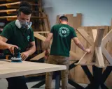 Un artisan porte un masque et utilise une ponceuse pour poncer une table en bois. Un autre artisan travaille sur un pied de table en bois.