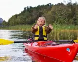 Glad kvinna i kajak med all kajak utrustning, ger tummen upp. Vacker natur med vass och träd i bakgrunden.