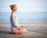 Kvinna med slutna ögon sitter i meditationsställning på en träbrygga vid havet, utövar progressiv avslappning.