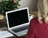Un ordinateur portable ouvert sur un bureau en bois, avec une tasse de café, des roses et un croissant. Le branchement PC est prêt pour le travail.