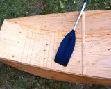 Bateau en contreplaqué brut, prêt pour la résine époxy. Une pagaie noire repose à l'intérieur.