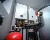 Dwa nowoczesne kotły gazowe wiszą na ścianie obok dużego, srebrnego bojlera. Pod nimi widoczny jest czerwony zbiornik przeponowy i skomplikowana instalacja hydrauliczna.